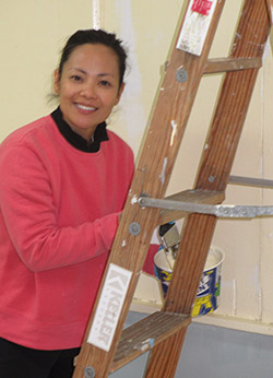 A woman standing next to a ladder while  holding a bucket of paint and a brush. she has a happy smile on her face.